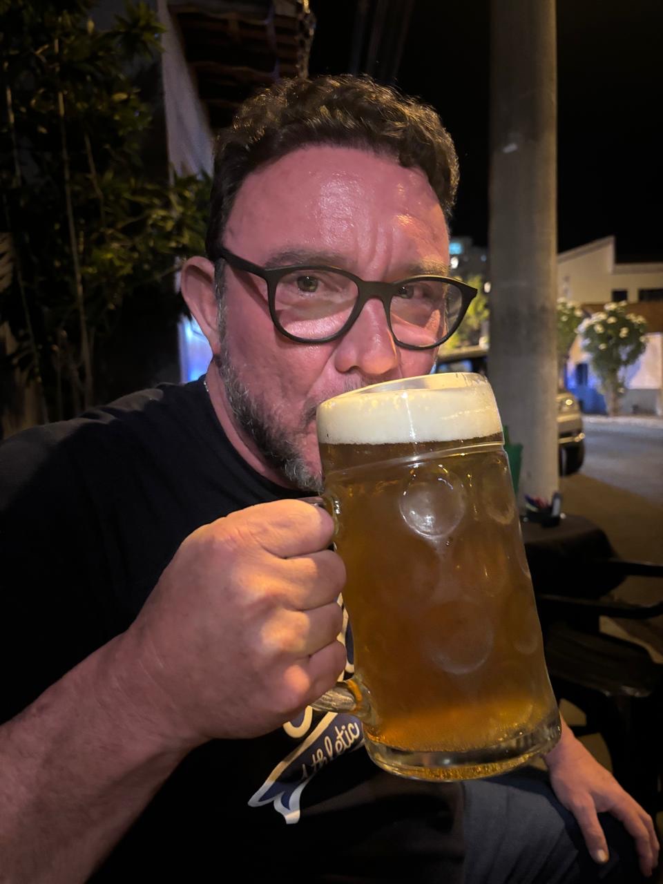 Dani erguendo uma caneca enorme de chopp, de óculos, à noite.