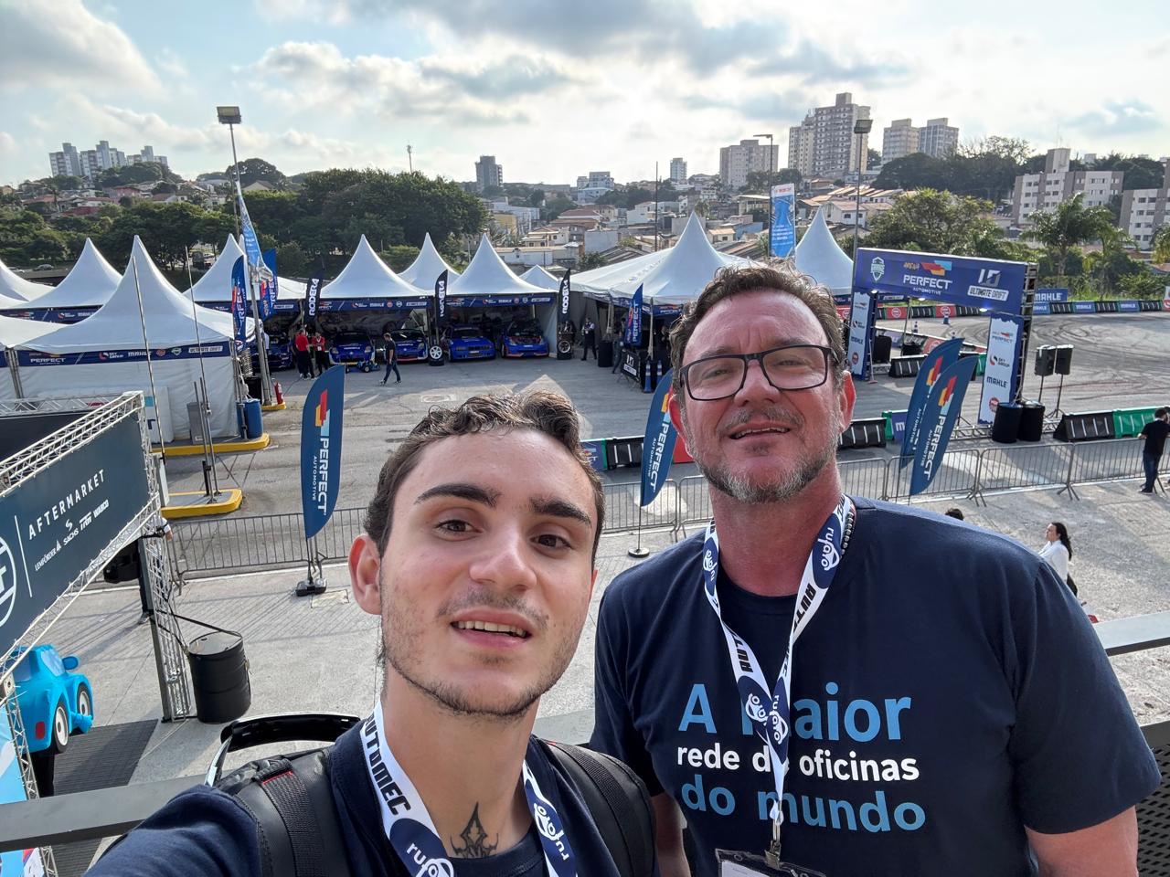 Dani e Felipe na feira Automec, tendas e carros ao fundo.
