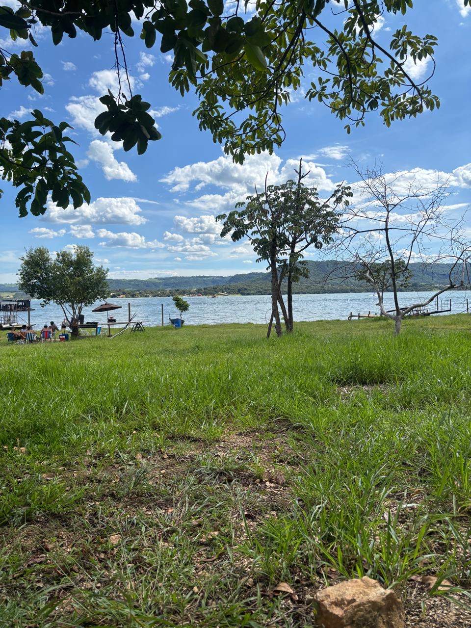 Gramado verde à beira da represa, com árvores e cadeiras de praia ao fundo.